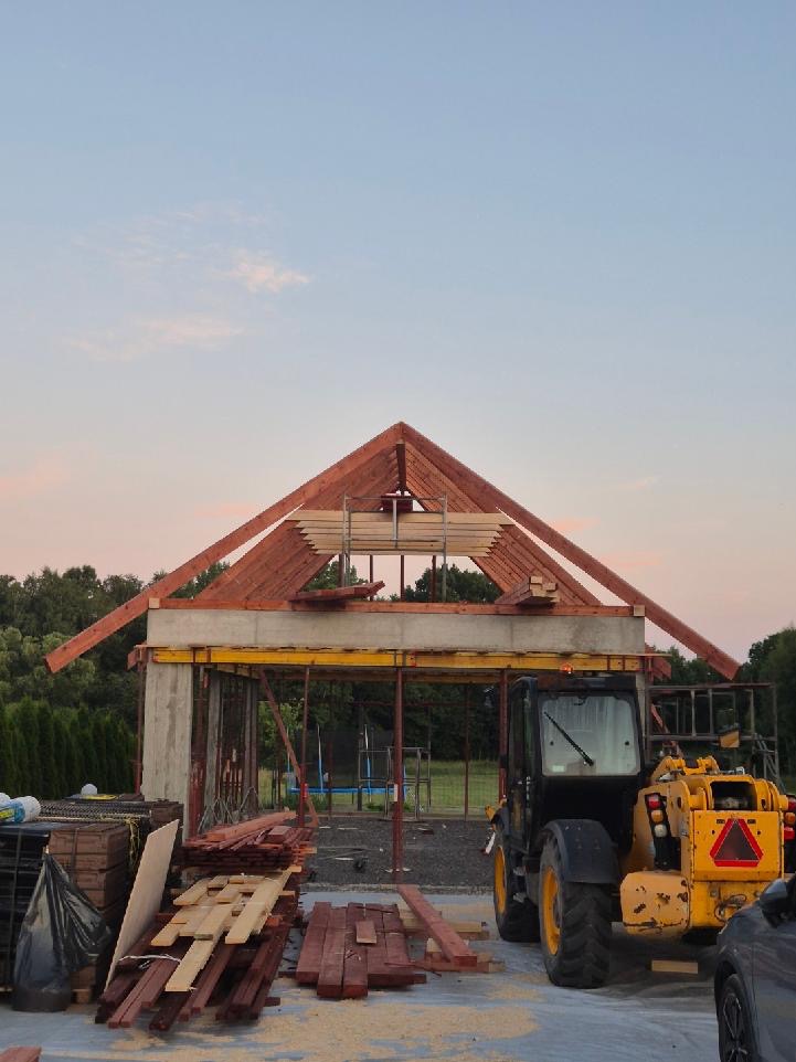 Budowa domu w trakcie: betonowa konstrukcja, drewniany szkielet dachu, deski, żółta ładowarka teleskopowa. Widok na las i niebo o zmierzchu.
