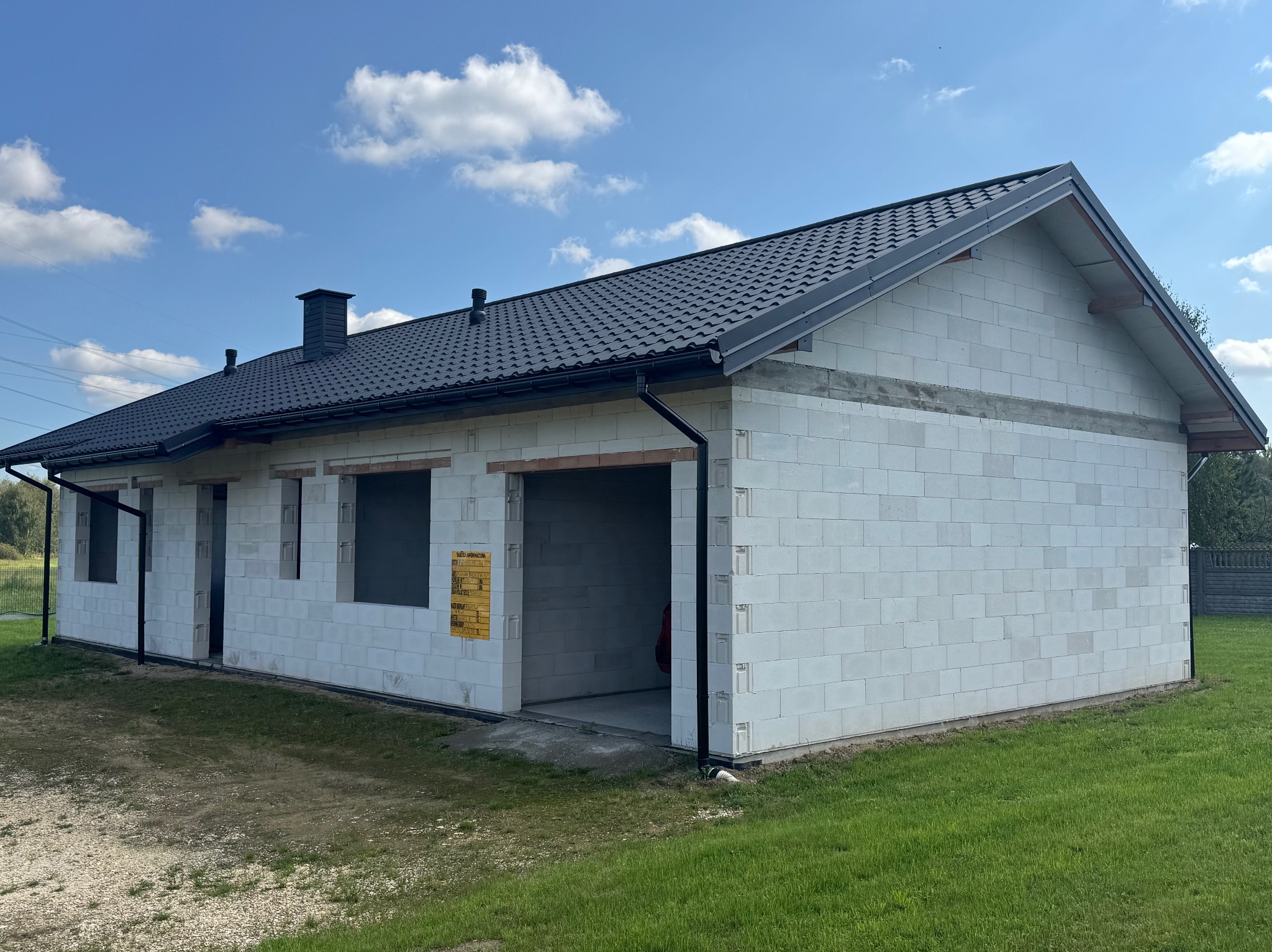 Niedokończony dom z białych bloczków, czarny dach z blachodachówki, widoczne okna i brama garażowa, zielona trawa, błękitne niebo. Budowa w toku.