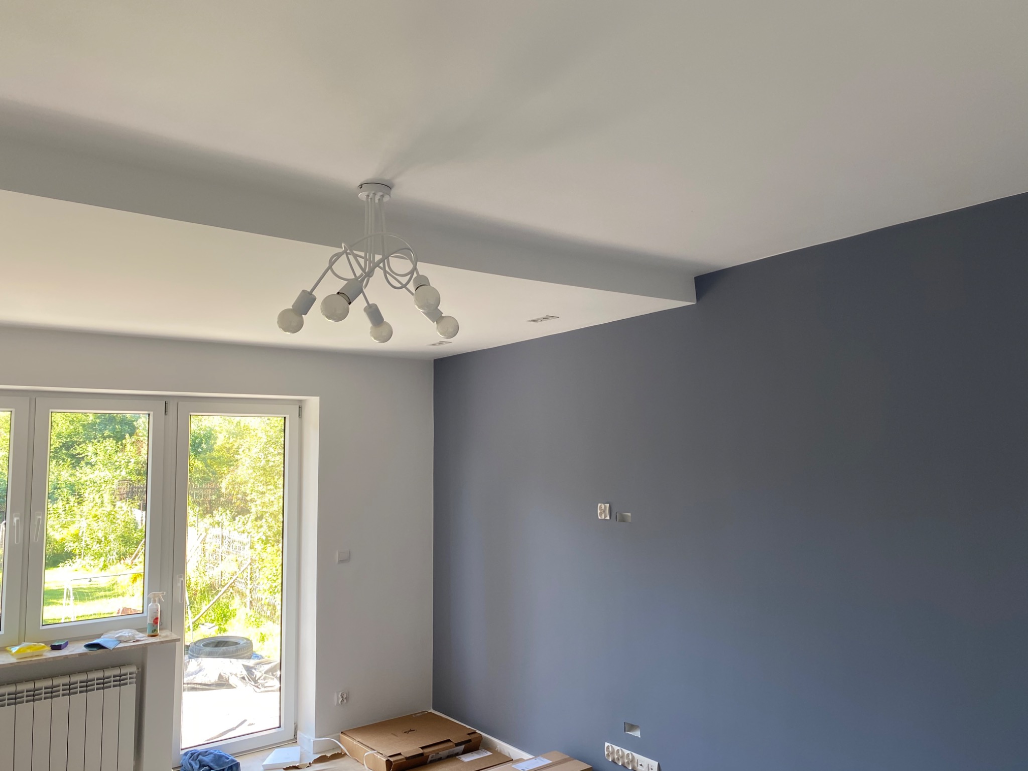 Pomieszczenie po malowaniu ścian na biało i szaro-niebiesko, widoczne kartony na podłodze, biały grzejnik pod oknem i nowoczesna lampa na suficie.