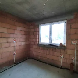 Surowy pokój z ceglanymi ścianami, widoczne rozprowadzone przewody elektryczne i puszki instalacyjne, okno z widokiem na zewnątrz, betonowy sufit.