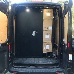 Rapido - Wnętrze busa z otwartymi drzwiami, zabezpieczone czarną metalową przegrodą zamykaną na klucz. Obok przegrody ustawione kartony z opisami i symbolami transportowymi.