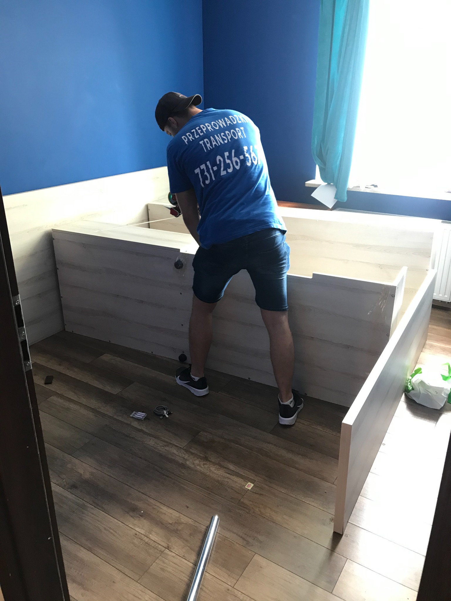 Mężczyzna w koszulce firmy transportowej skręca mebel w pokoju z niebieską ścianą, widoczne narzędzia i elementy montażowe na podłodze.