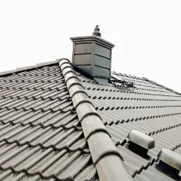 Szary dach z ceramicznej dachówki karpiówki, widoczny komin z szarym wykończeniem i odpowietrzniki dachowe. Widok z dołu pod kątem.