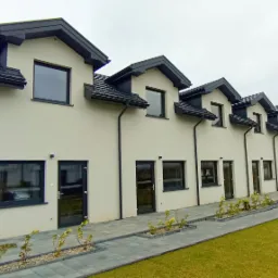 Szeregówka domów jednorodzinnych z ciemnymi dachami i rynnami, jasna elewacja, szara kostka brukowa, trawnik, pochmurne niebo.