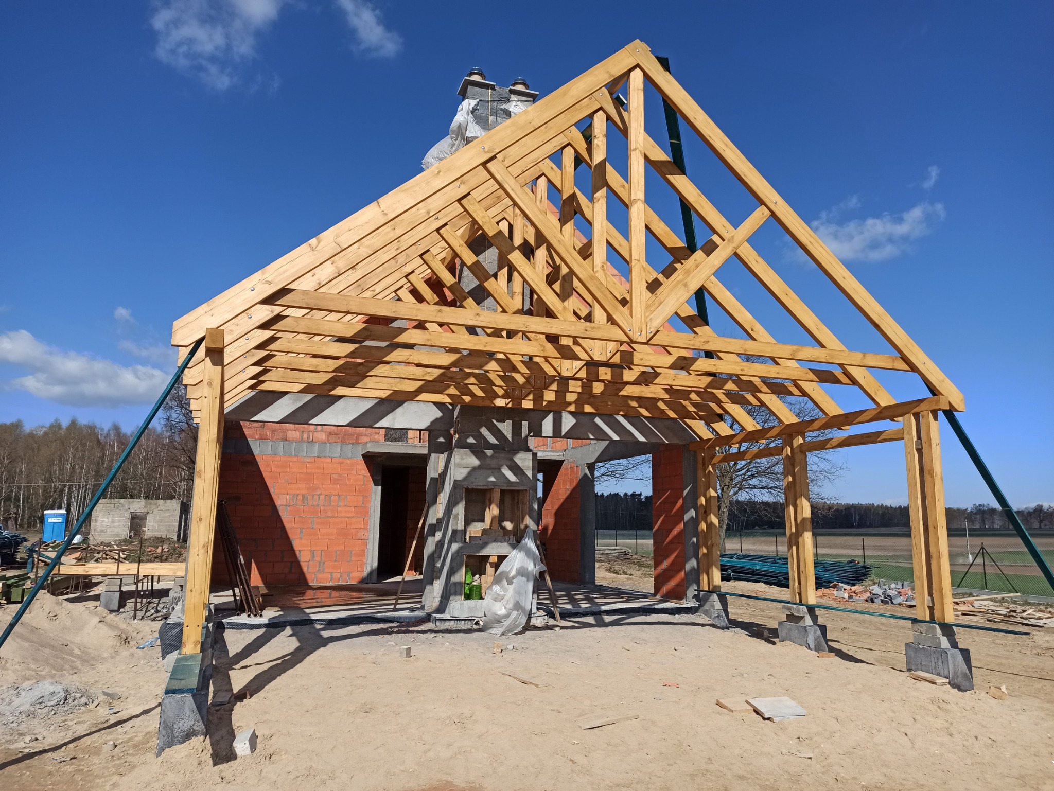 Konstrukcja drewniana więźby dachowej w trakcie budowy, widoczne ceglane ściany budynku, niebieskie niebo w tle.