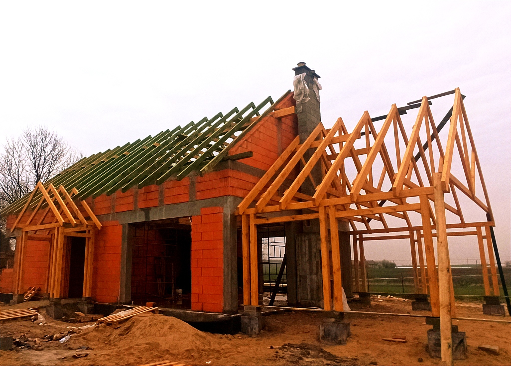 Budowa domu jednorodzinnego w stanie surowym otwartym z widoczną konstrukcją więźby dachowej z drewnianych belek, murowanym kominem zabezpieczonym folią i ścianami z czerwonej cegły, na tle...