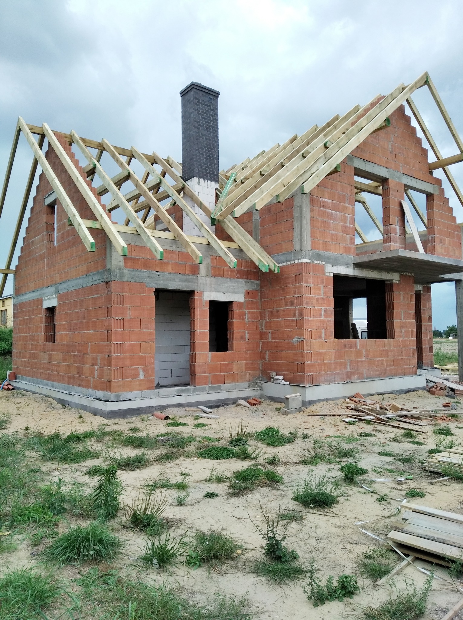 Surowy stan budowy domu jednorodzinnego z czerwonej cegły z widoczną konstrukcją drewnianą dachu i kominem z cegły klinkierowej na tle pochmurnego nieba.