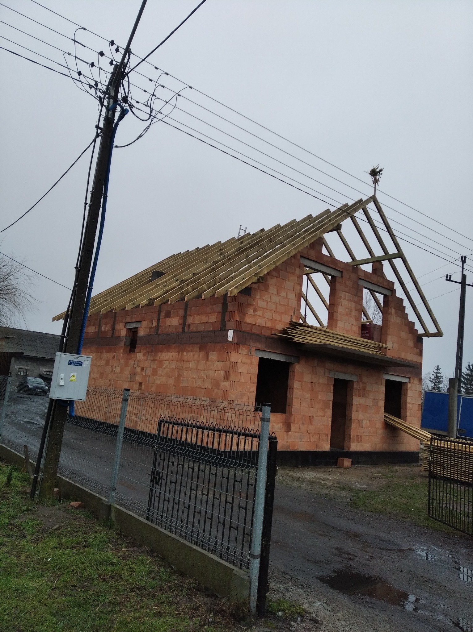 Surowy stan domu z czerwonej cegły z widoczną konstrukcją dachu z drewnianych łat, ozdobiony wieńcem na szczycie, na tle pochmurnego nieba i słupa energetycznego