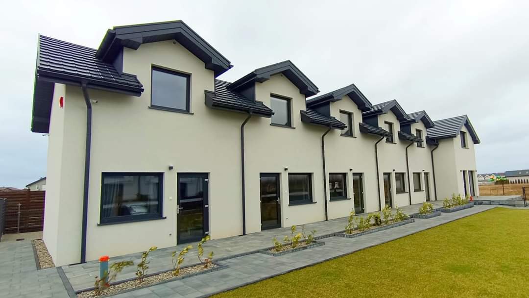 Szereg identycznych, dwukondygnacyjnych domów z ciemnoszarą dachówką i rynnami, jasnymi elewacjami, ciemnymi ramami okiennymi i drzwiami, oraz zagospodarowanym terenem z trawnikiem i chodnikiem...