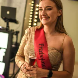 Uśmiechnięta kobieta w złotej sukience i szarfie z napisem 'Przebojowa Latka' trzyma kieliszek czerwonego wina, w tle oświetlone lustro i sprzęt DJ-ski.