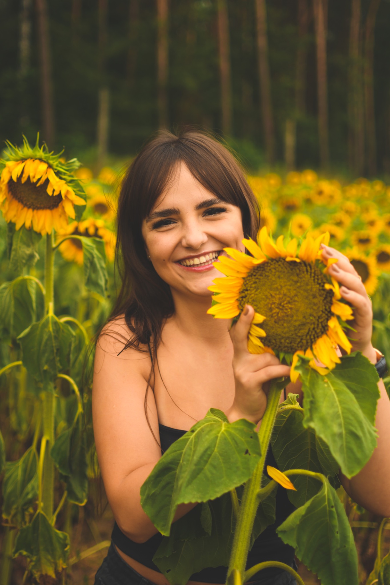 Uśmiechnięta brunetka na polu słoneczników trzyma duży kwiat słonecznika, widoczne brązowe włosy z grzywką i fragment czarnego topu.