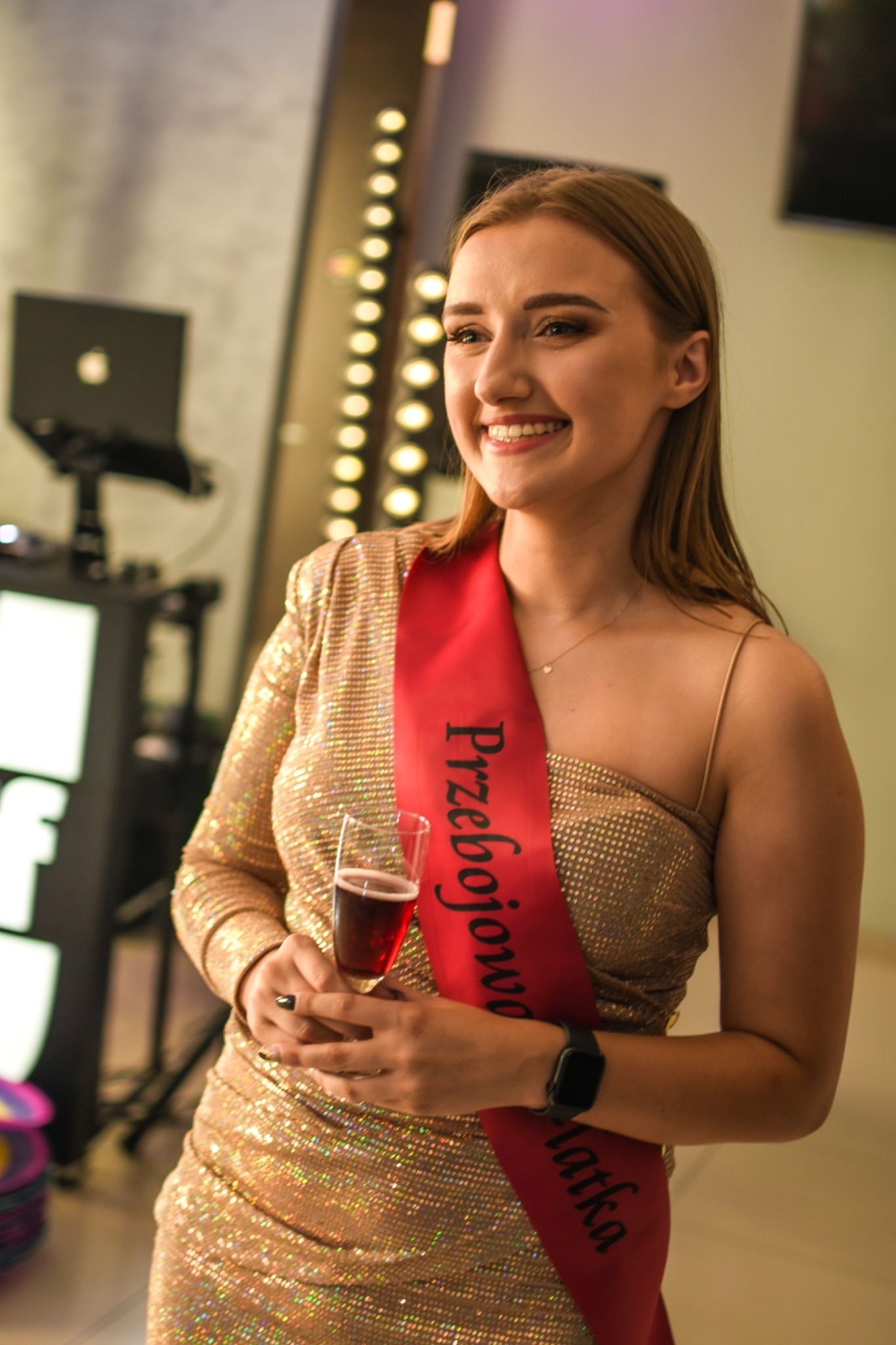 Uśmiechnięta kobieta w złotej sukience i szarfie z napisem 'Przebojowa Latka' trzyma kieliszek czerwonego wina, w tle oświetlone lustro i sprzęt DJ-ski.