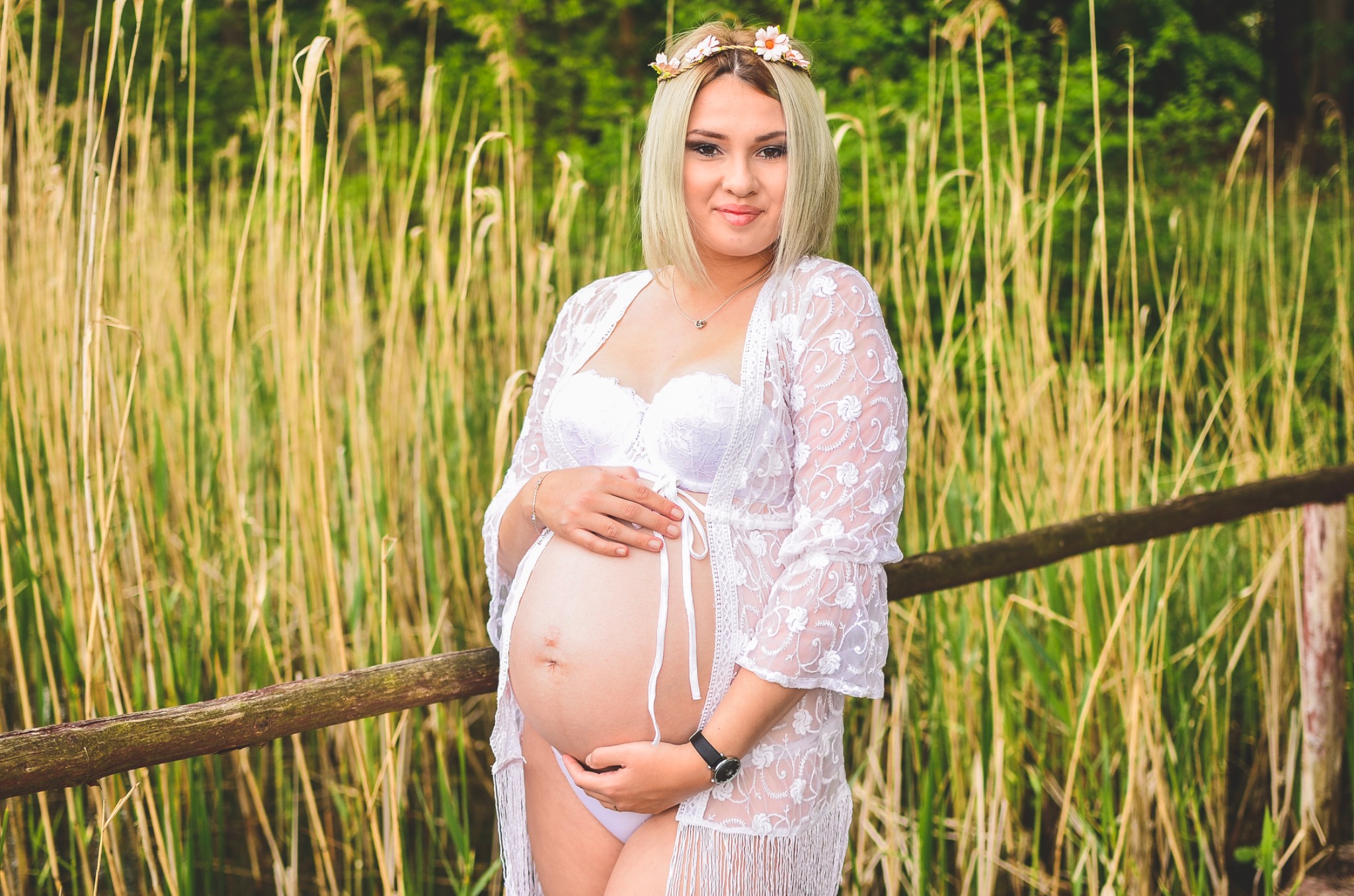 Kobieta w ciąży w białej bieliźnie i narzutce z koronkowym wzorem, pozująca na tle trzcin i drewnianego płotku, z wiankiem z kwiatów na głowie.