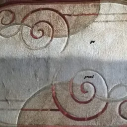 Owalny dywan z wzorem spiralnym, widoczna różnica w czystości: górna połowa wyczyszczona, dolna zabrudzona, obok leżą akcesoria do czyszczenia.