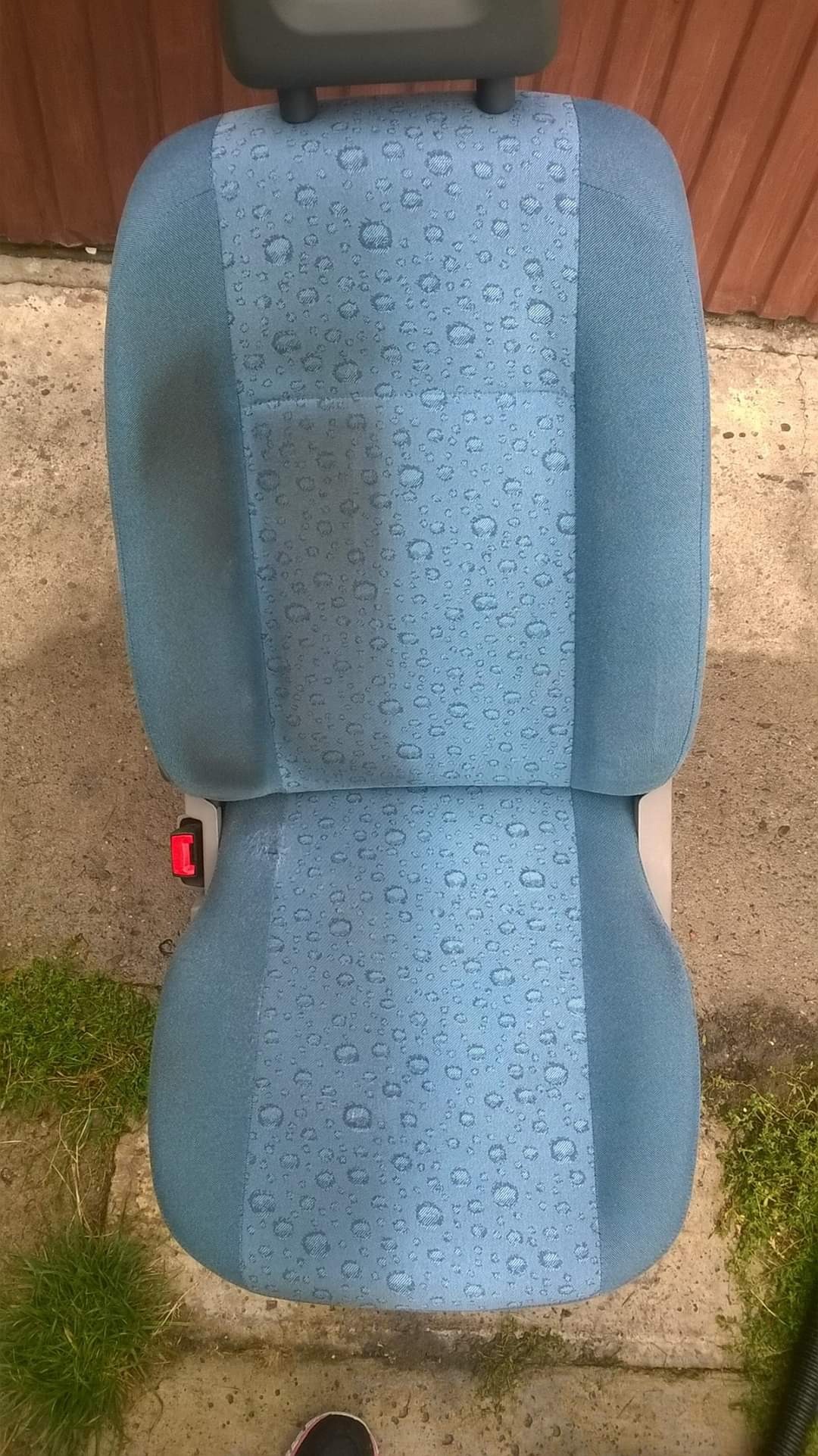 Wyjęty z samochodu, niebieski fotel samochodowy z wzorem kropelek, leżący na betonowym podłożu obok trawy i drewnianego płotu.