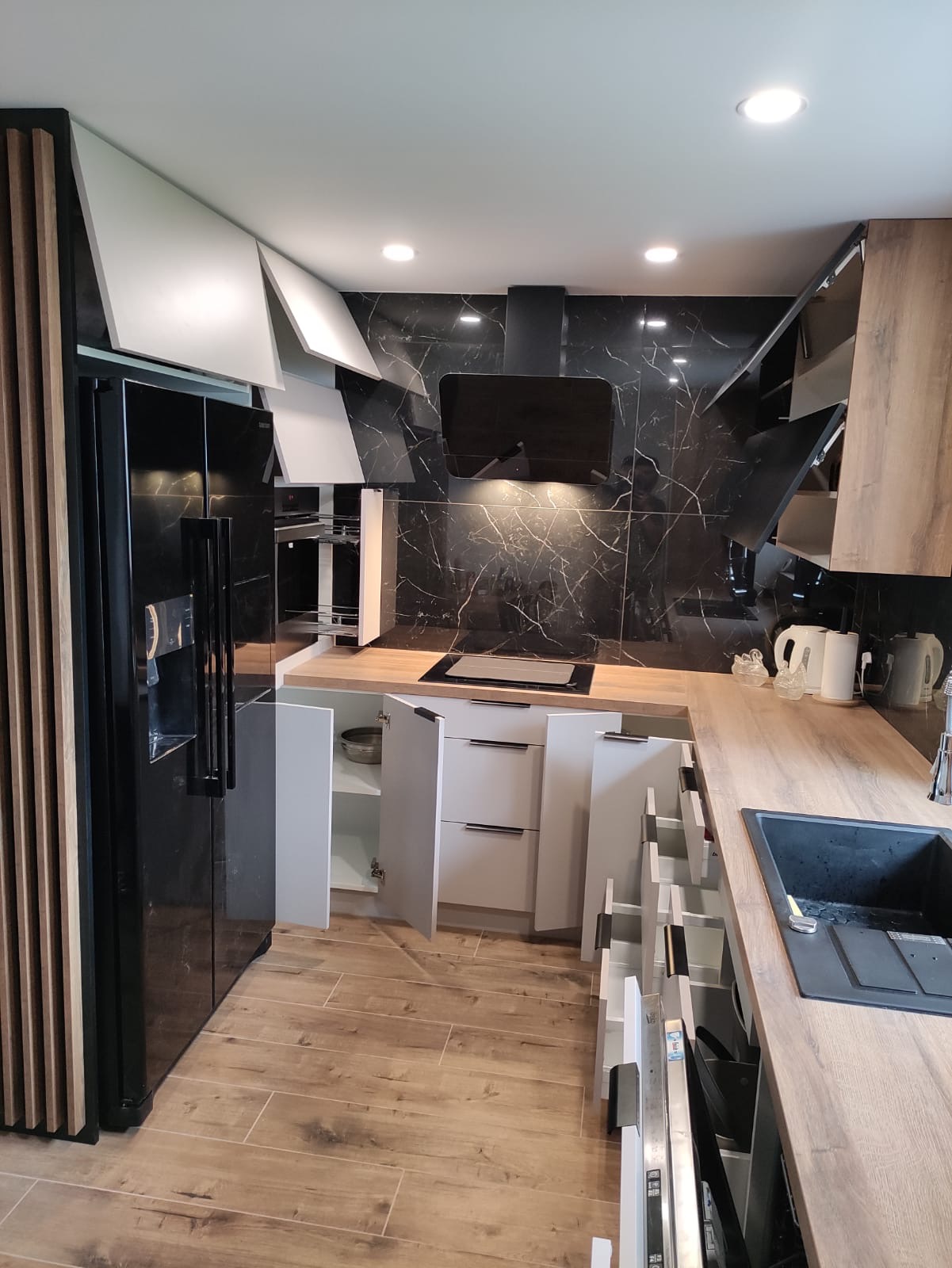 Nowoczesna kuchnia z czarnymi marmurkowymi płytkami na ścianie, drewnianym blatem, białymi szafkami z otwartymi drzwiczkami i szufladami, czarną lodówką i drewnianą ścianką działową.