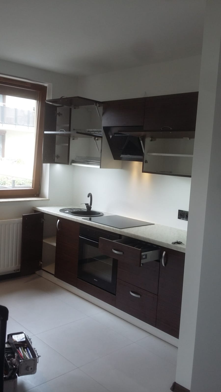 Ciemnobrązowa zabudowa kuchenna z jasnym blatem, czarnym zlewem i płytą indukcyjną. Otwarta szafka górna z suszarką na naczynia oraz wysunięta szuflada na sztućce. W tle widoczne okno i skrzynka...