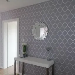 Szara ściana z wzorem marokańskiej koniczyny, okrągłe lustro z dekoracyjną ramą, biała konsola z wazonem i srebrną figurką, widok na białe drzwi i fragment podłogi