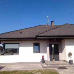 Nowoczesny dom jednorodzinny z białą elewacją, ciemną dachówką i antracytowymi oknami, z kwiatami w doniczkach przy wejściu i pod oknami, antena na dachu.