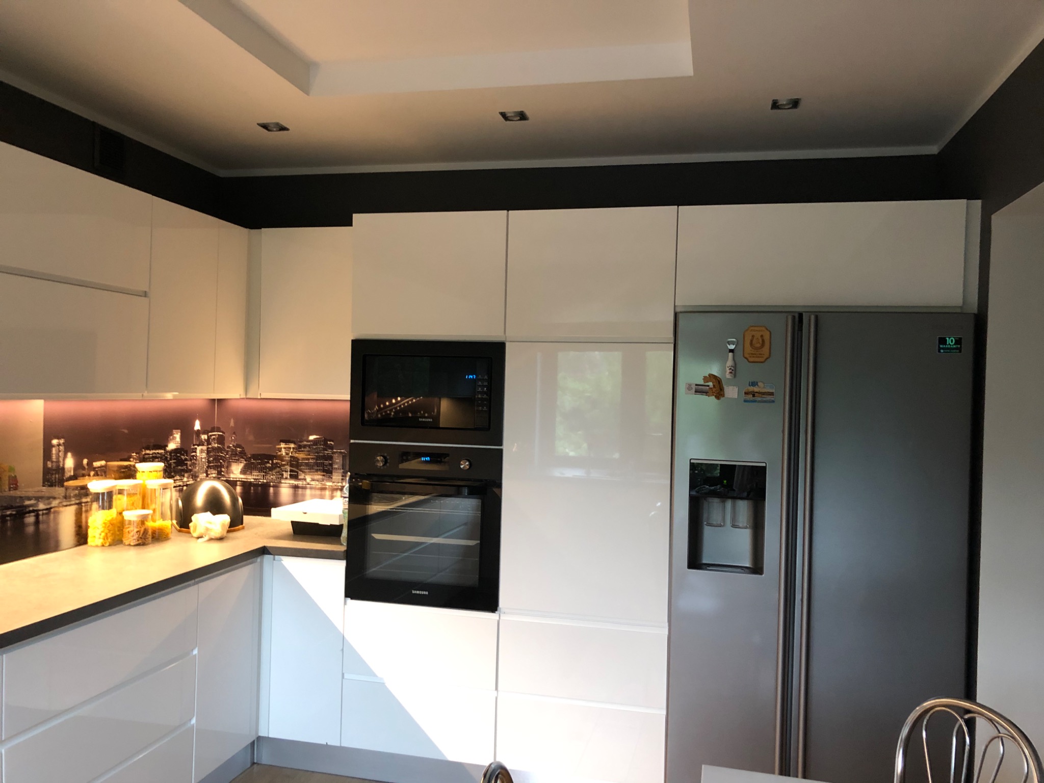 Nowoczesna, jasna kuchnia z białymi szafkami, szarym blatem, piekarnikiem i lodówką side-by-side, z czarnym pasem pod sufitem i fototapetą z motywem miasta nad blatem.