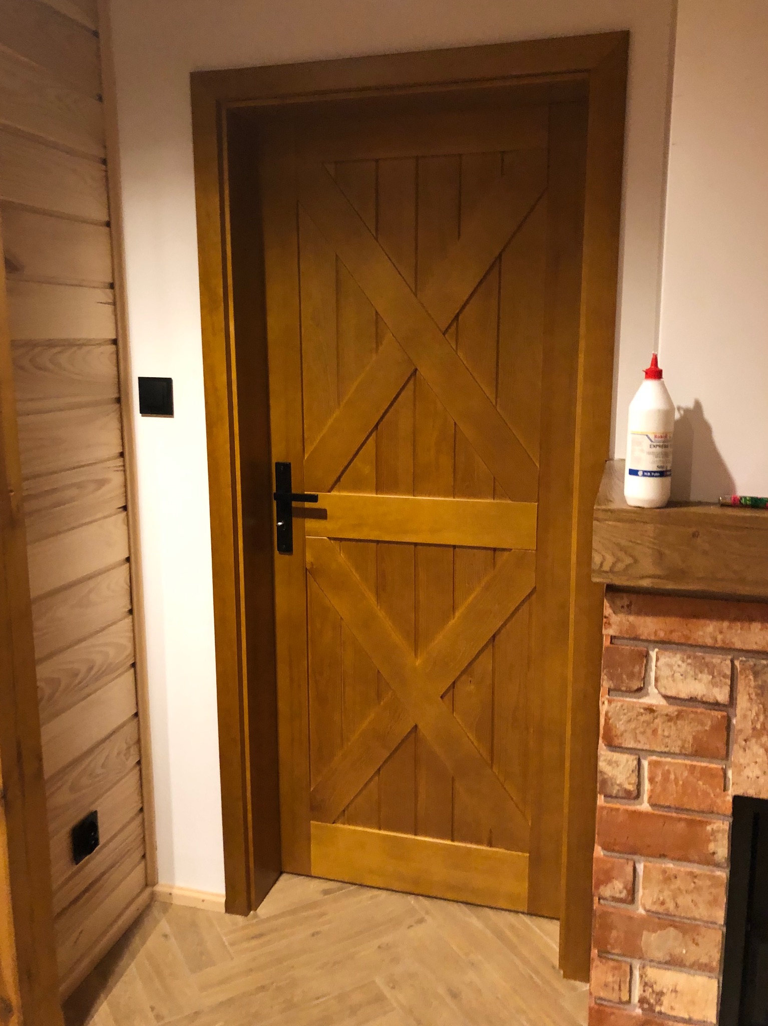 Drewniane drzwi wewnętrzne w stylu rustykalnym z czarną klamką, widoczne obok ściany z boazerią i fragmentu ceglanego kominka z butelką kleju na półce.