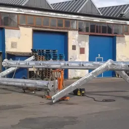 Dwie duże, srebrne, metalowe konstrukcje, prawdopodobnie elementy hali, leżą na zewnątrz budynku z niebieskimi bramami. Widoczny wózek transportowy, palety i spawarka.