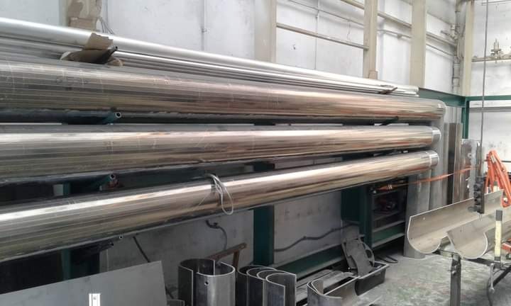 Stalowe rury ułożone na metalowym regale w hali produkcyjnej, widoczne także elementy konstrukcyjne i narzędzia.