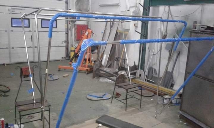 Metalowa konstrukcja z rur, owinięta niebieską folią ochronną, w warsztacie z narzędziami i sprzętem, prawdopodobnie w trakcie spawania lub montażu.