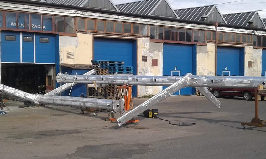 Dwie duże, srebrne, metalowe konstrukcje, prawdopodobnie elementy hali, leżą na zewnątrz budynku z niebieskimi bramami. Widoczny wózek transportowy, palety i spawarka.