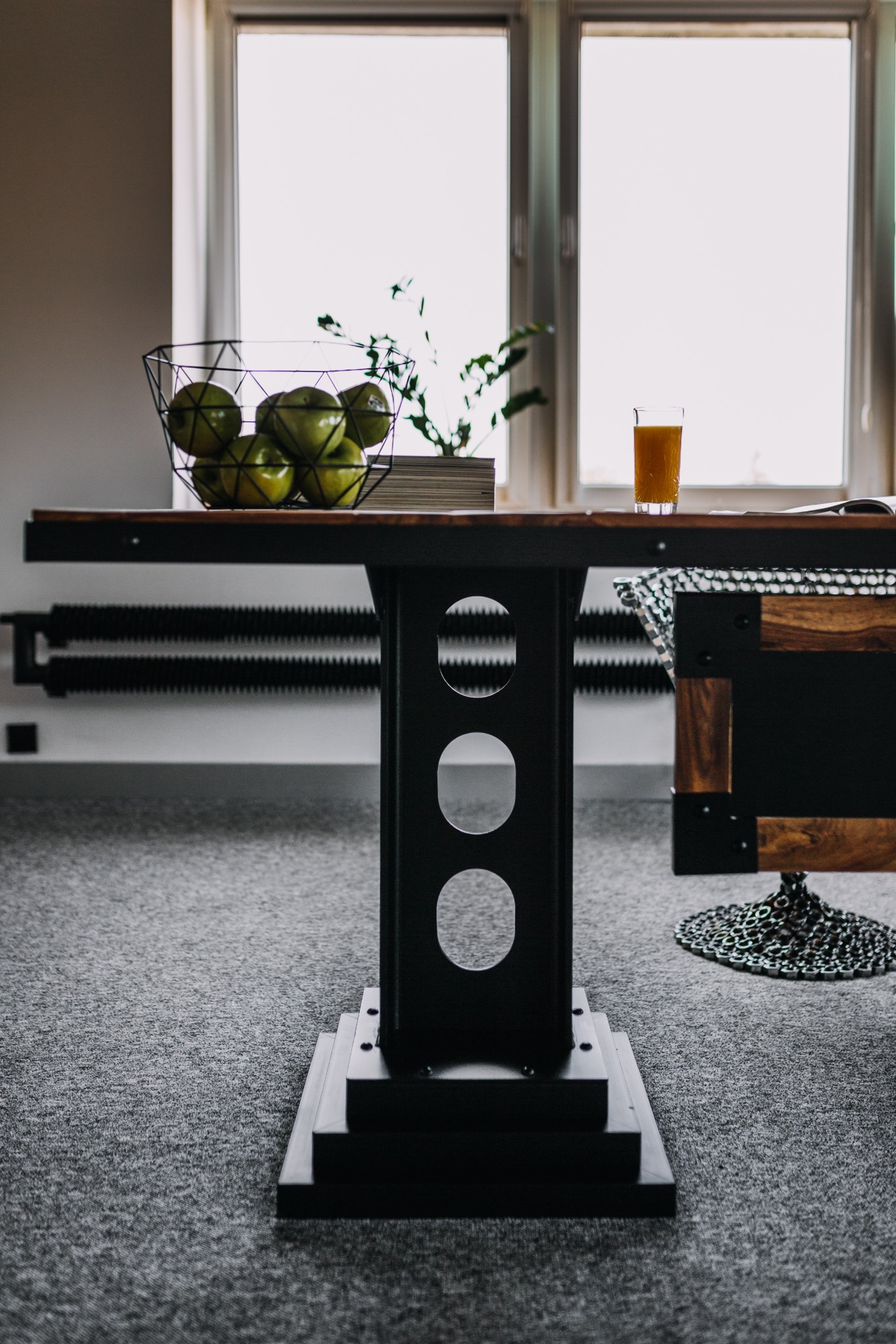 Stół z drewnianym blatem i metalową podstawą z okrągłymi otworami, na którym stoi koszyk z zielonymi jabłkami i szklanka soku, widok na okno w tle.