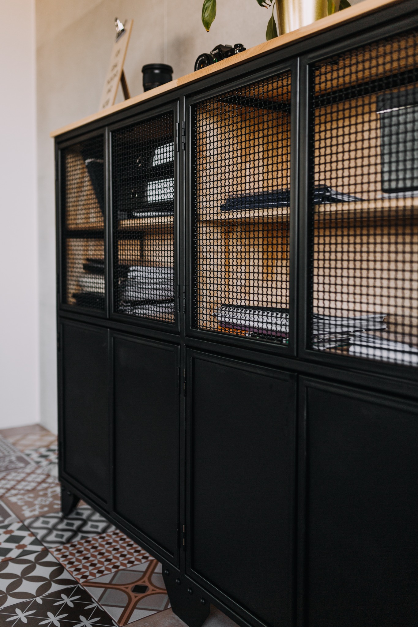 Czarna, metalowa komoda z siatkowymi drzwiczkami, drewnianym blatem, stojąca na wzorzystej podłodze, z widocznymi książkami i akcesoriami w środku.