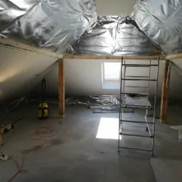 Poddasze w trakcie adaptacji: ocieplenie folią paroizolacyjną, częściowo zamontowane płyty kartonowo-gipsowe, drabina, lampa budowlana i odkurzacz na betonowej podłodze.