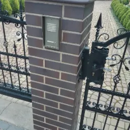 Murowany słupek ogrodzeniowy z domofonem i dekoracyjnym, kutym metalowym ogrodzeniem w kolorze czarnym, prowadzącym do domu z brukowaną ścieżką.