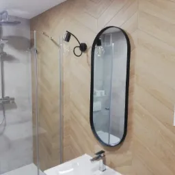 Nowoczesna łazienka z kabiną prysznicową typu walk-in, umywalką z chromowaną baterią i owalnym lustrem w czarnej ramie, oświetlona lampą z żarówkami Edisona, wykończona płytkami imitującymi drewno...