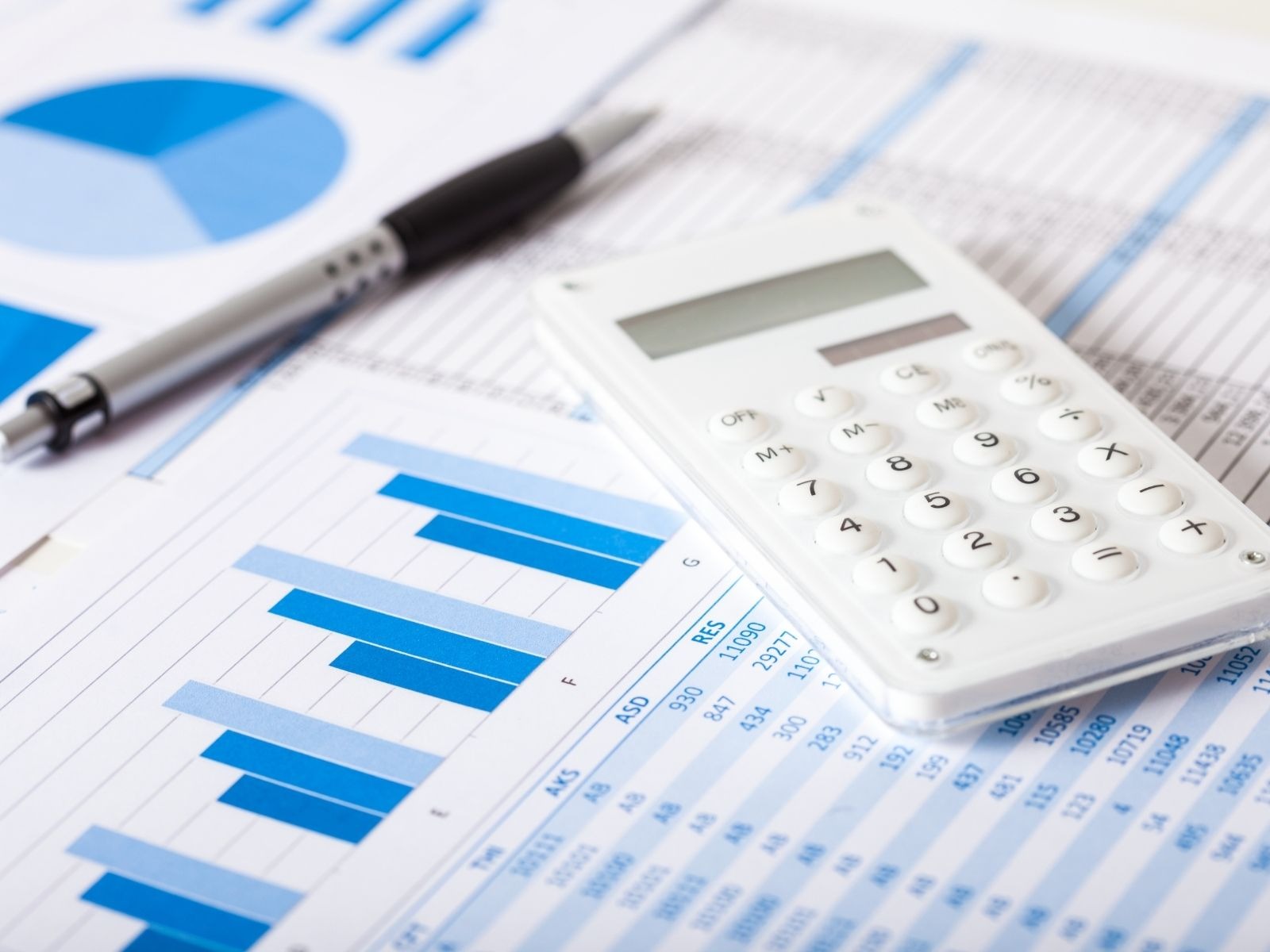 Biały kalkulator, czarny długopis i arkusze z niebieskimi wykresami słupkowymi i tabelami danych finansowych.