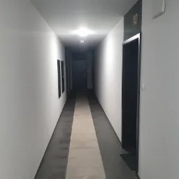 Długi korytarz w nowoczesnym budynku z białymi ścianami, szarą podłogą i ciemnymi drzwiami z numerem 10, świeżo po malowaniu.