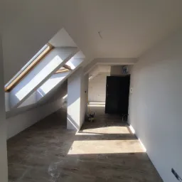 Poddasze po remoncie z oknami dachowymi, świeżo pomalowane na biało, z brązową podłogą imitującą drewno i czarnymi drzwiami; widoczne ślady po pracach wykończeniowych.