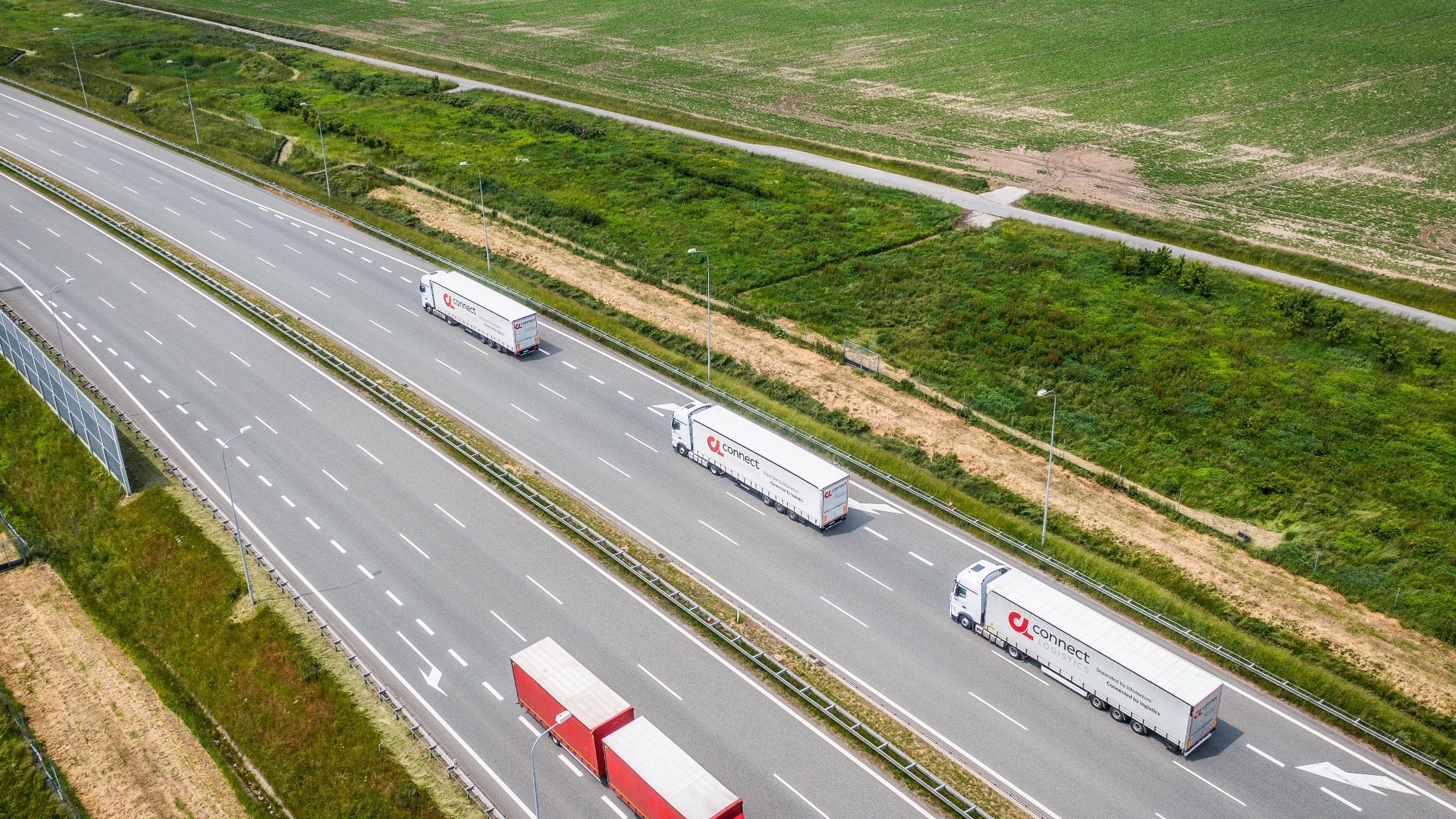 Widok z góry na autostradę z trzema białymi ciężarówkami z logo 'connect logistics' i jedną czerwoną, jadącymi w różnych kierunkach, otoczoną zielonymi polami.