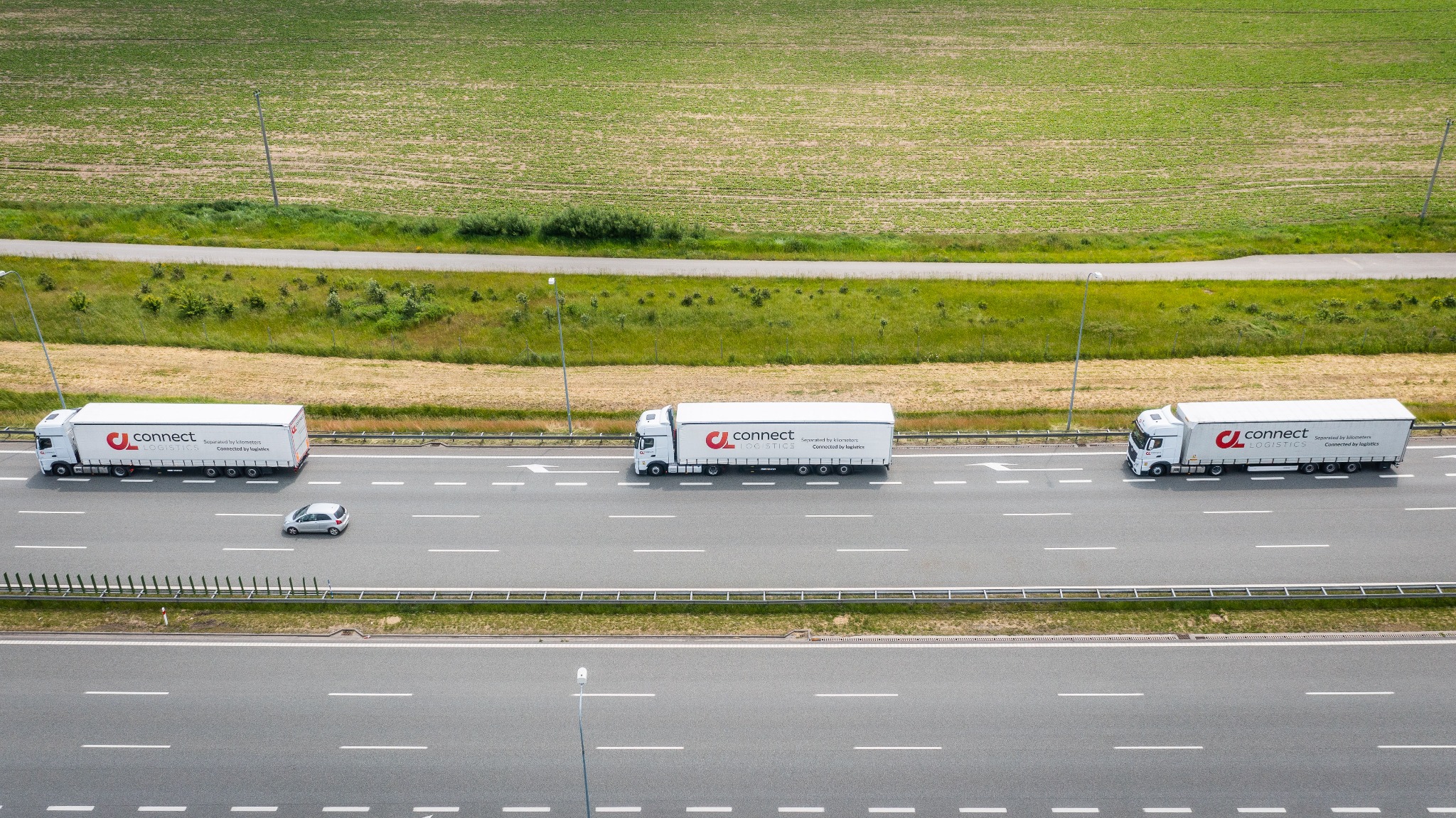 Trzy białe ciężarówki z logo Connect Logistics jadą autostradą w kierunku horyzontu, widziane z góry, z polem w tle i szarym samochodem osobowym na pasie obok.