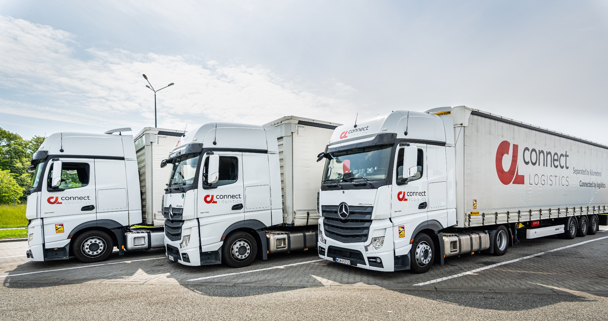 Trzy białe ciężarówki z naczepami firmy Connect Logistics zaparkowane na placu, widok z przodu i boku, z widocznym logo firmy na naczepach.