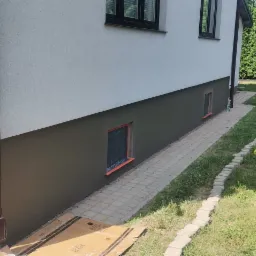 Fragment elewacji budynku po częściowym pomalowaniu, dolna część w kolorze grafitowym, okna zabezpieczone folią, na trawniku leżą żółte kubki i radio.