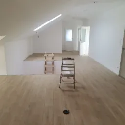 Poddasze po remoncie z nową podłogą z paneli, białymi ścianami, oknem dachowym i dwiema parami drzwi, drabina i schodki w tle.