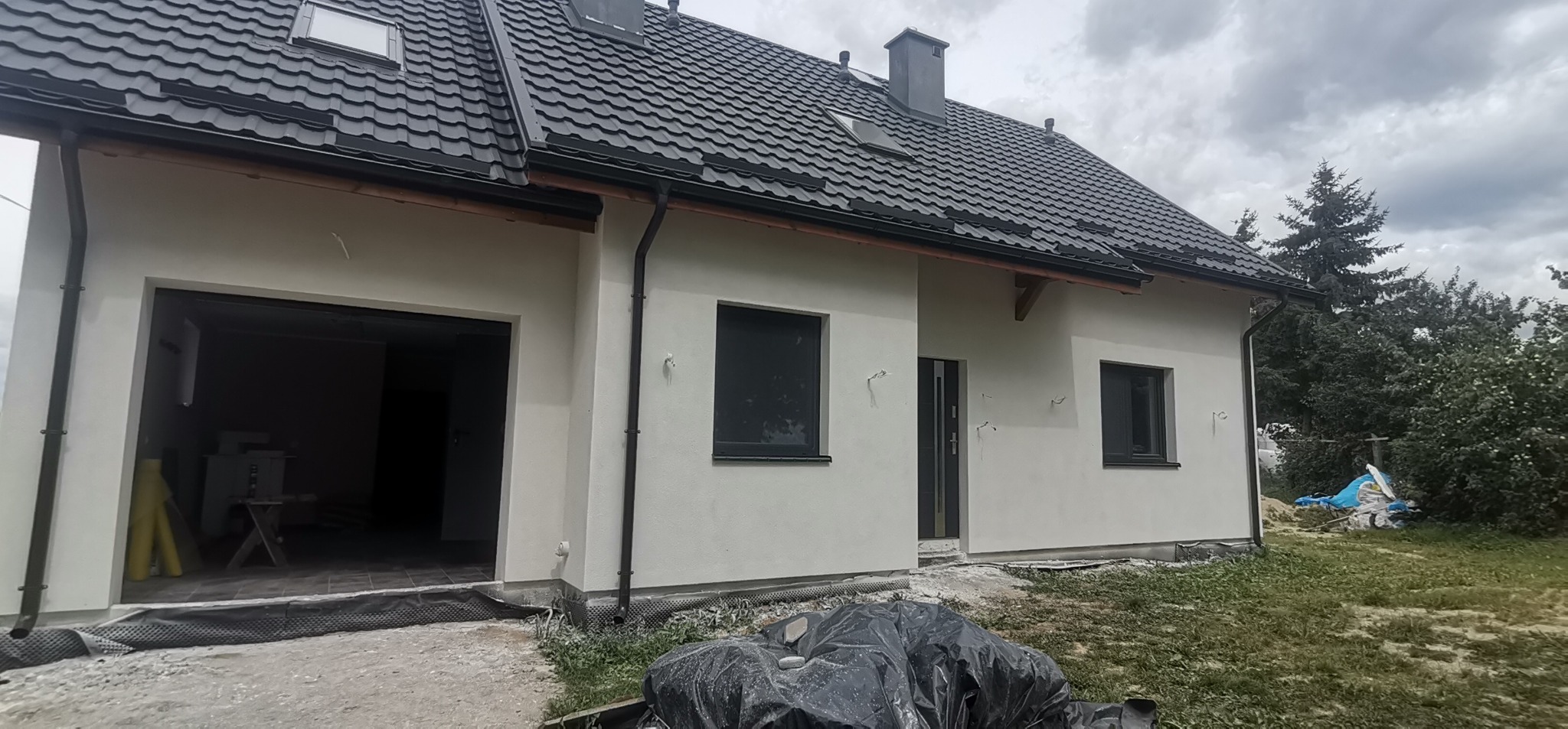 Nowo wykończony dom jednorodzinny z szarą elewacją, czarnymi oknami i dachem z blachodachówki, widoczne niedokończone prace wykończeniowe wokół budynku.