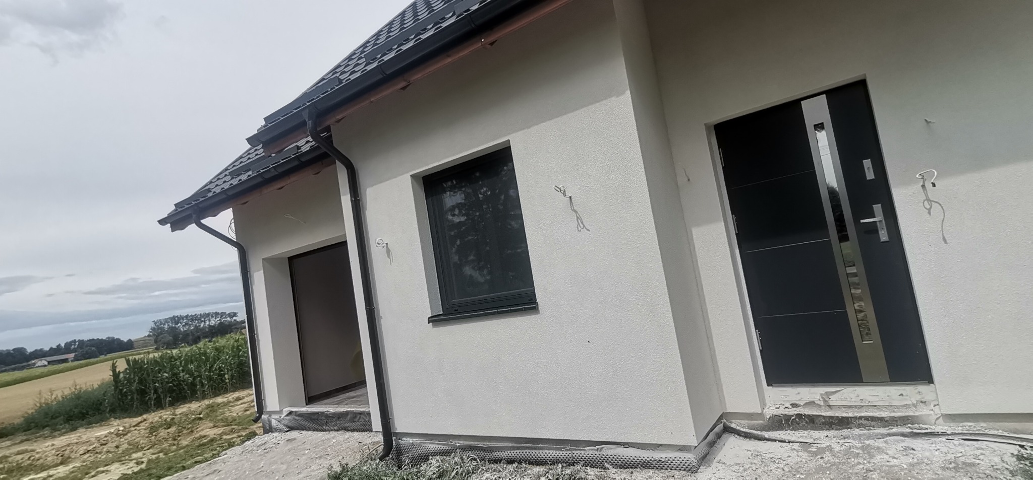 Fragment elewacji domu jednorodzinnego z szarą stolarką okienną i drzwiami wejściowymi, widoczne niedokończone prace wykończeniowe przy cokole.