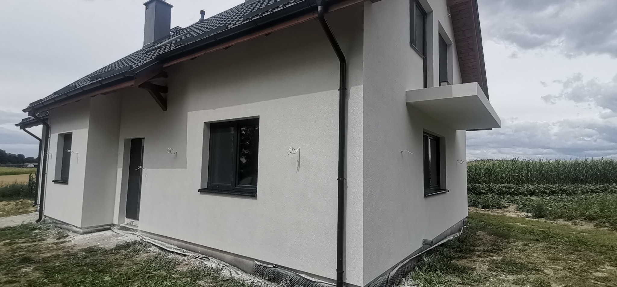 Świeżo otynkowany dom jednorodzinny z szarą stolarką okienną i czarną dachówką, widok z boku na tle pola kukurydzy i pochmurnego nieba.