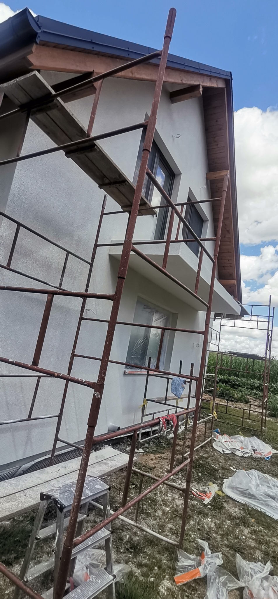 Boczny widok domu w trakcie odnawiania elewacji, z rusztowaniem, zabezpieczonymi oknami folią i widocznymi śladami farby na ziemi.