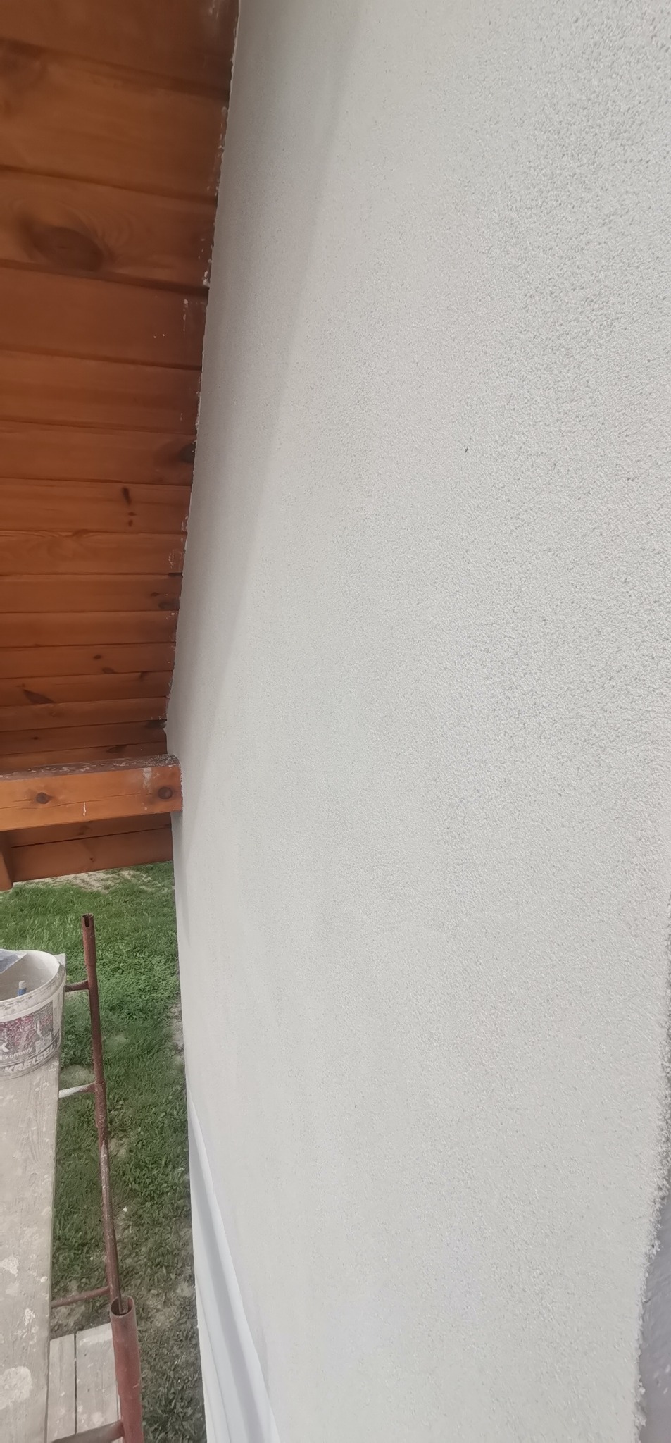 Świeżo otynkowana ściana budynku z widocznym fragmentem drewnianej konstrukcji dachu i rusztowaniem z wiadrem farby u dołu.