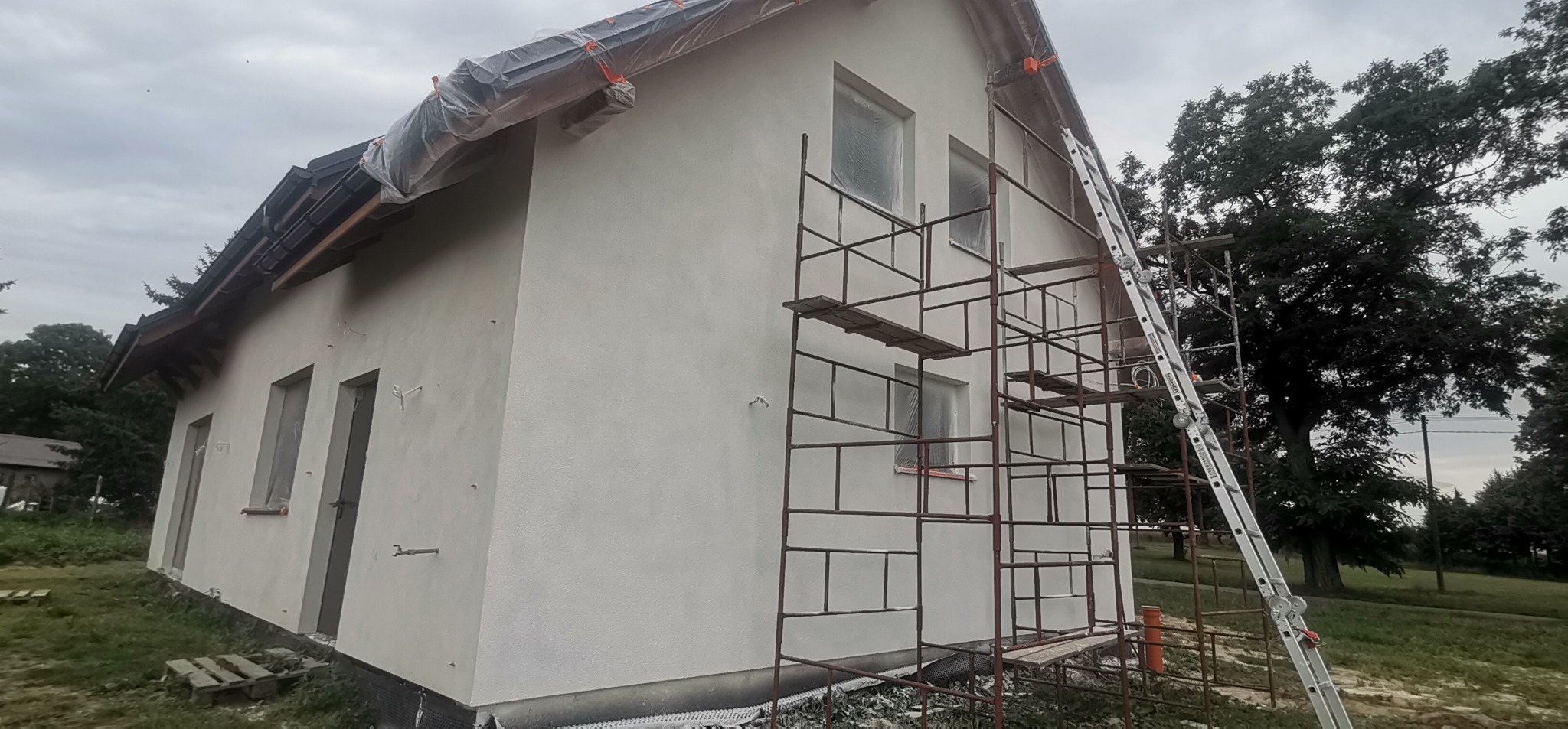 Dom jednorodzinny z nową, jasną elewacją w trakcie prac wykończeniowych; okna i dach zabezpieczone folią, widoczne rusztowania i drabina.