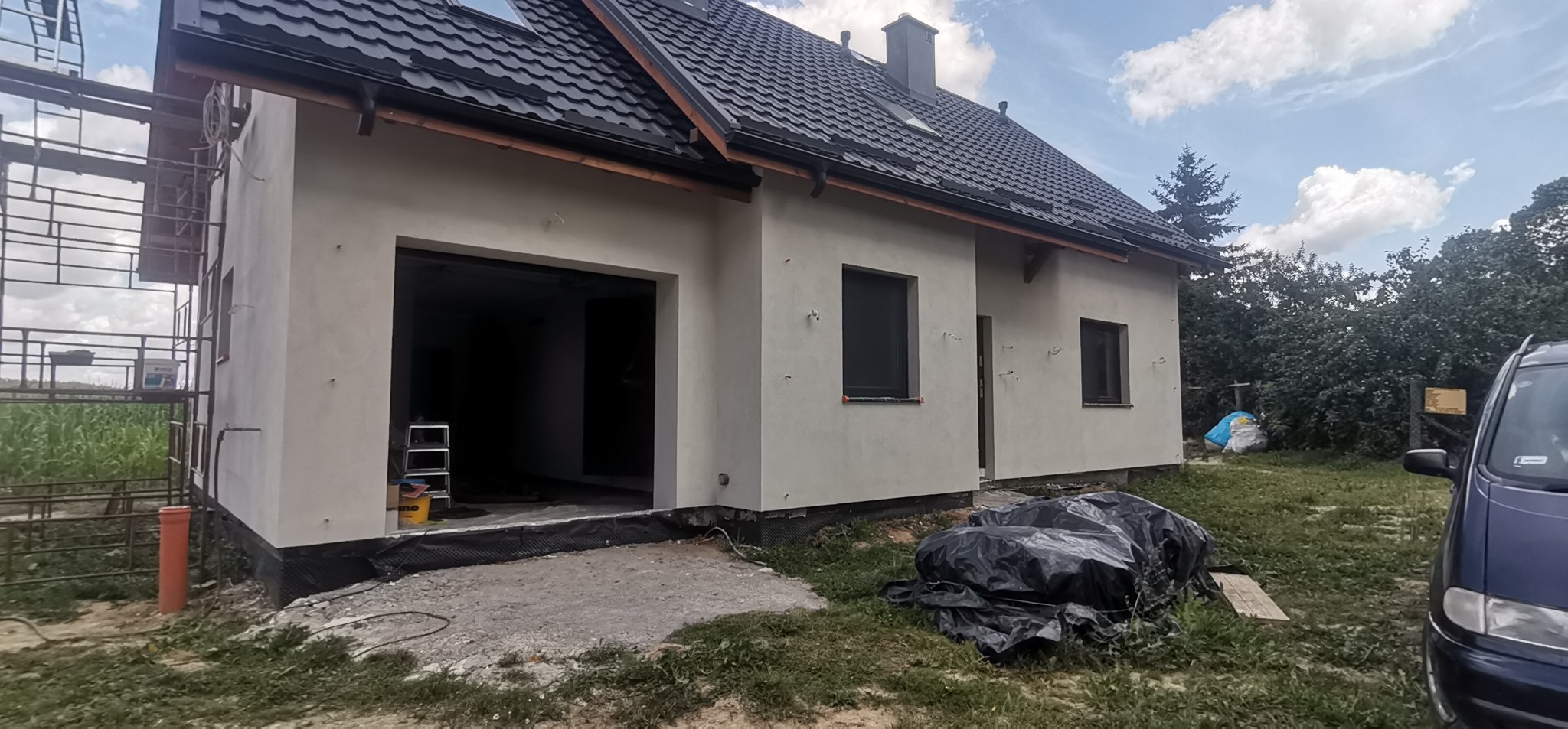 Dom jednorodzinny w trakcie prac elewacyjnych: surowa, jasna elewacja, czarny dach, widoczne rusztowania, drabina w otwartym garażu, oraz zabezpieczone folią materiały budowlane na trawniku.