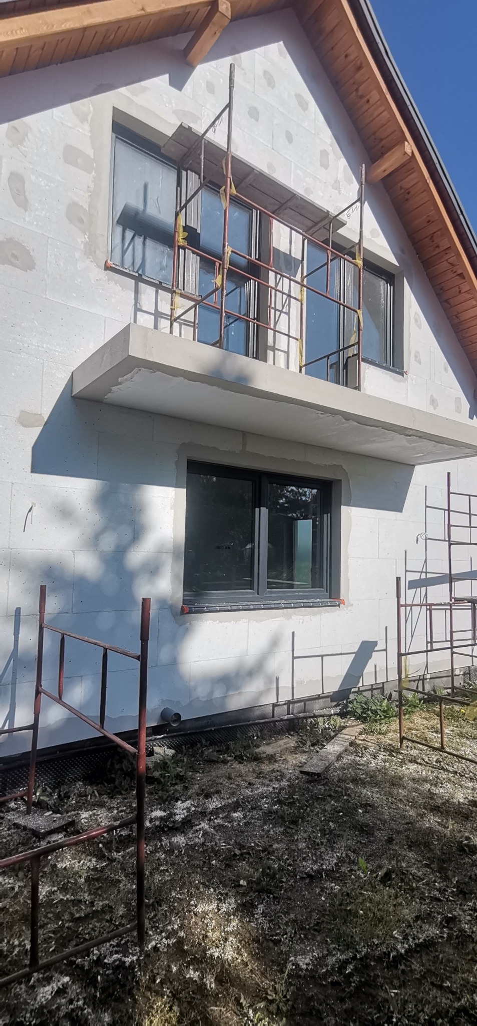 Surowa elewacja budynku w trakcie budowy z widocznymi rusztowaniami i oknami w kolorze antracytowym.